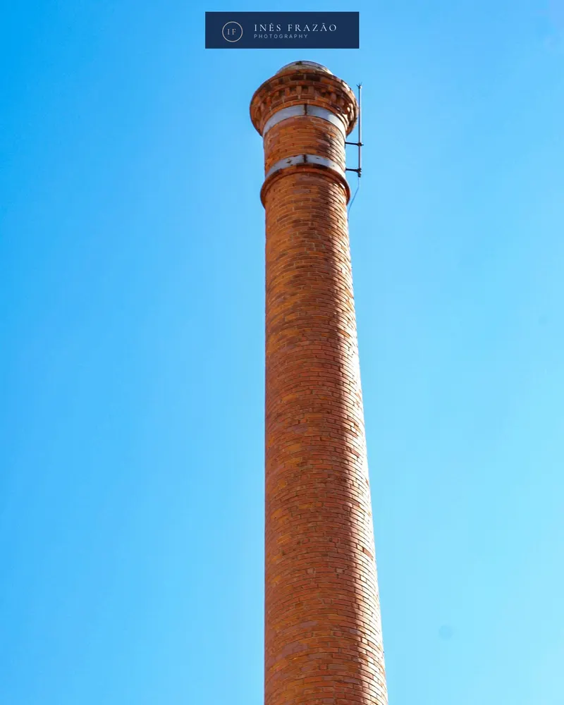 Chaminé industrial de tijolo a erguer-se contra um céu azul limpo, fotografia de arquitetura por Inês Frazão.