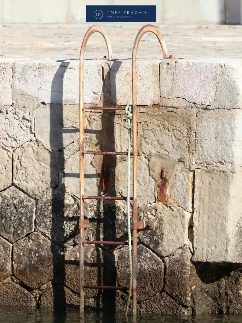 Escada de ferro enferrujada presa a um muro de pedra junto à água, fotografia de pormenor arquitetónico por Inês Frazão.