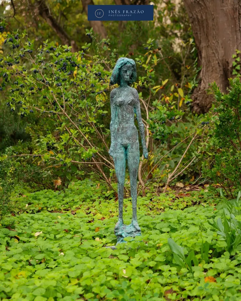 Estátua de bronze de uma figura feminina rodeada de vegetação verde num jardim, fotografia de pormenor urbano por Inês Frazão.