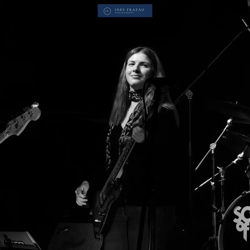 Baixista sorridente a tocar em palco, a preto e branco, fotografia de concerto por Inês Frazão.