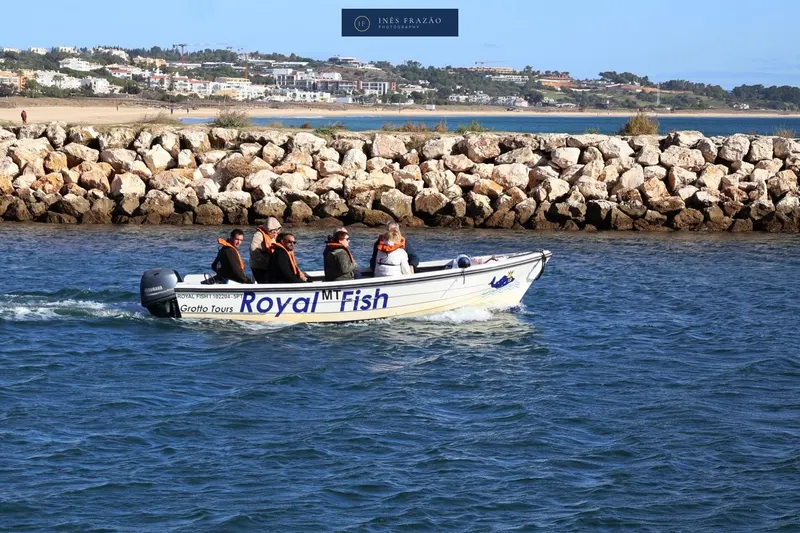 Pequeno barco com passageiros a navegar junto a um quebra-mar de pedra, fotografia de paisagem costeira por Inês Frazão.