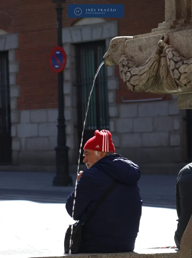 Homem de gorro vermelho sentado numa rua junto a uma fonte, fotografia de retrato de rua por Inês Frazão.