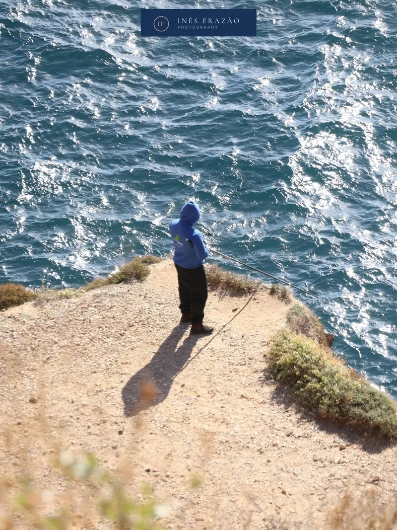 Retrato distante de um pescador à beira da falésia, captando a solidão tranquila e a imensidão do mar. Fotografia documental por Inês Frazão.