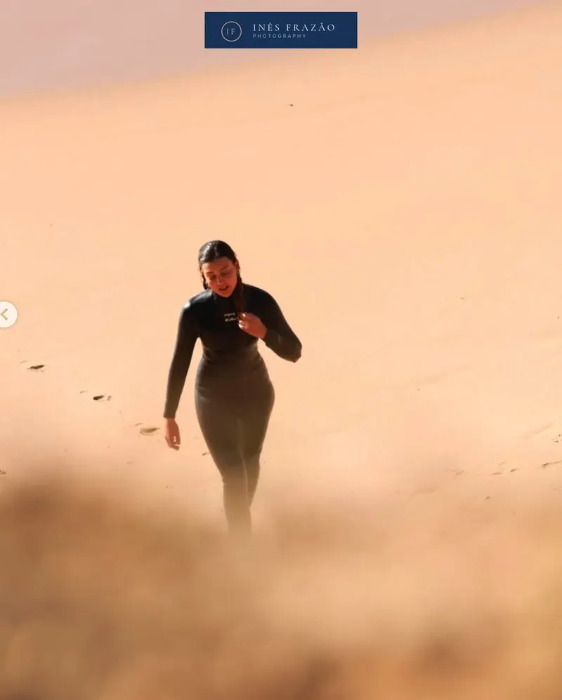 Retrato espontâneo de uma surfista a subir a praia, iluminada pela luz suave e envolvida pelas texturas quentes da areia. Fotografia documental por Inês Frazão.